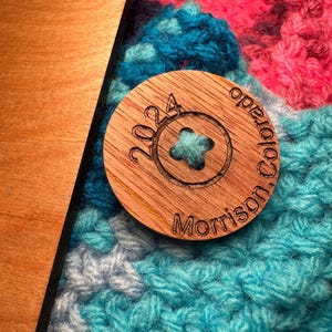 Close-up of a round, wooden button featuring the text '2024 Morrison Colorado' attached to a crochet blanket. The button is resting on a teal, pink, and gray crocheted fabric. The button is made of light brown wood with dark brown engraved text. The crocheted fabric has a textured surface. The button is approximately 1.5 inches in diameter.