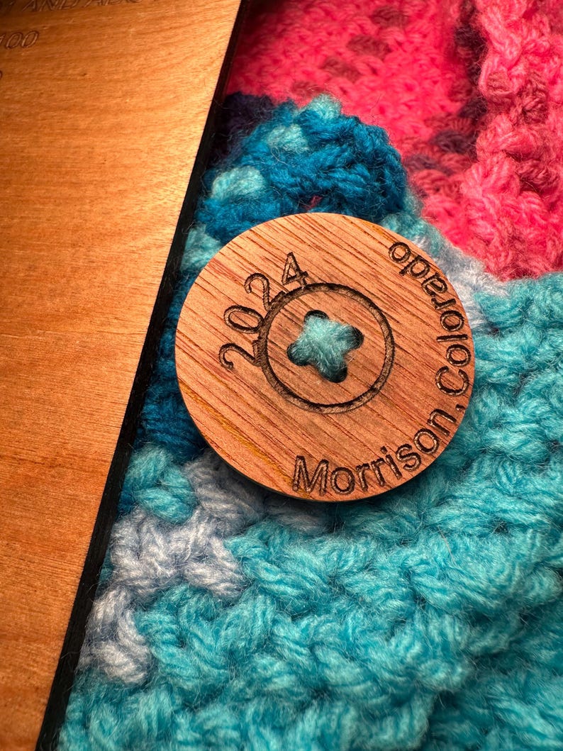 Close-up of a round, wooden button featuring the text '2024 Morrison Colorado' attached to a crochet blanket. The button is resting on a teal, pink, and gray crocheted fabric. The button is made of light brown wood with dark brown engraved text. The crocheted fabric has a textured surface. The button is approximately 1.5 inches in diameter.