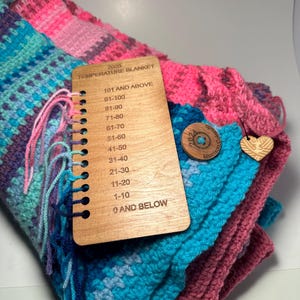 Close-up of a folded crocheted temperature blanket in shades of pink, teal, and purple.  A wooden temperature key is attached, listing temperature ranges from 0 and below to 101 and above. The blanket is handmade and features a color-coded design corresponding to the temperature key.  The key is labeled '2025 Temperature Blanket'.
