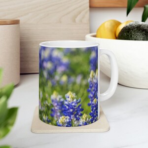 Könnte beinhalten: Weiße Keramiktasse mit einem bunten Bild von blauen Lupinen. Die Tasse hat einen Henkel auf der rechten Seite.