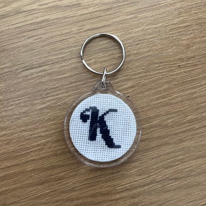 May include: A white and black cross-stitch keychain with the letter K in a clear plastic frame.
