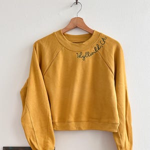 Puede incluir: Sudadera corta de color amarillo mostaza con la inscripción bordada "Idyllwild, CA". La sudadera está colgada de una percha de madera. Una ilustración de cangrejo enmarcada, un pañuelo marrón y un sombrero beige están sobre una superficie de madera.