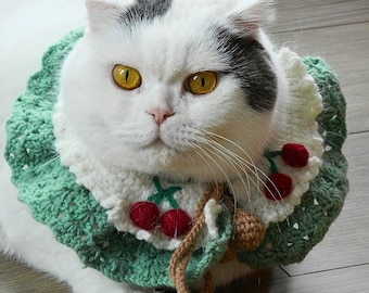 May include: A white cat with a black spot on its head wearing a green and white crocheted collar adorned with red cherry accents. The cat has yellow eyes and is sitting on a wooden floor.