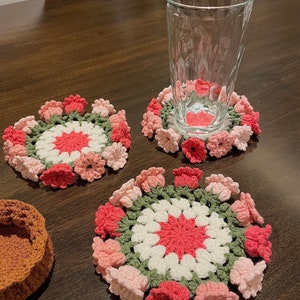 May include: Four crocheted coasters with a floral design in shades of pink, white, and green. The coasters are round and have a raised centre. A glass is sitting on one of the coasters.
