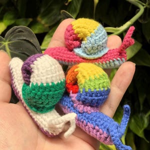 May include: Three crocheted snails in rainbow colors. The snails are made with different colored yarn, including purple, green, white, blue, yellow, orange, and red.
