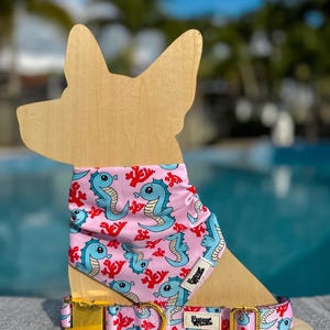 Könnte beinhalten: Halsband und Bandana-Set für Hunde mit einem Seepferdchen- und Korallenmuster in Hellrosa, Blau und Rot. Das Halsband hat eine goldfarbene Schnalle und einen passenden Seepferdchen-Druck. Das Bandana ist um eine Holz-Hunde-Silhouette gelegt.
