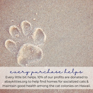 May include: A paw print in the sand with the text "every purchase helps" and the text "Every little bit helps. 10% of our profits are donated to abaykitties.org to help find homes for socialized cats & maintain good health among the cat colonies on Hawaii."  The text is in a white box with a logo of a cat in a white circle with the text "A Bay Kitties" and a logo of a book with the text "Dinny O'Lance Sue".