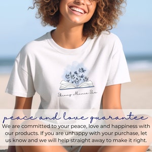 May include: A white t-shirt with a blue and white illustration of an open book with flowers on top. The text "Skinny Minnie Sue" is written below the book. The t-shirt is worn by a person with curly hair, smiling, sitting on a beach.