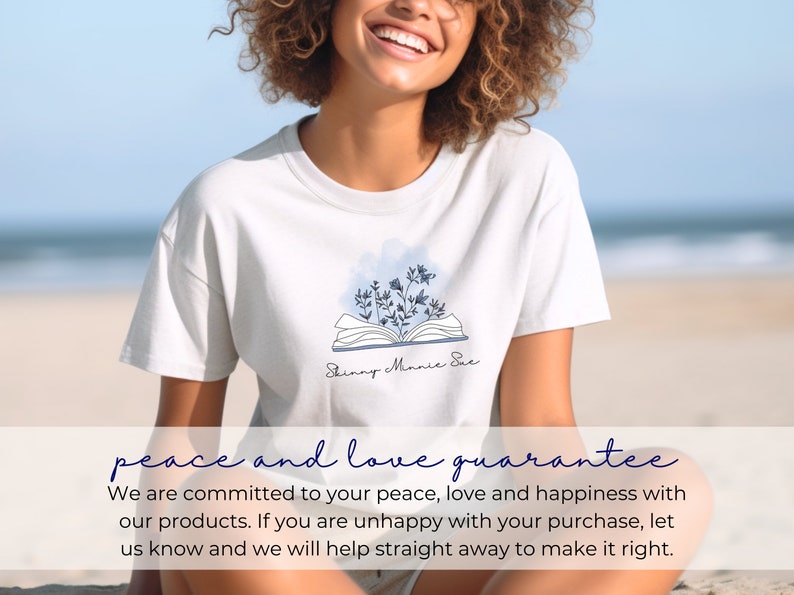 May include: A white t-shirt with a blue and white illustration of an open book with flowers on top. The text "Skinny Minnie Sue" is written below the book. The t-shirt is worn by a person with curly hair, smiling, sitting on a beach.