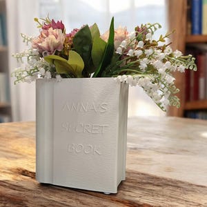 May include: White ceramic book-shaped vase with the text "Anna's Secret Book" embossed on the front, filled with pink and white flowers.