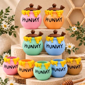 May include: A collection of nine round honey pots in various pastel colors including pink, orange, teal, blue, and tan. Each pot features the word HUNNY printed in black, with sculpted yellow honey drips around the rim and a brown lid on top.