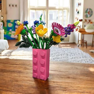 Può includere: Un vivace bouquet di fiori colorati in un vaso rosa in stile mattone. I fiori includono fiori gialli, rosa, blu e viola con steli e foglie verdi. Il vaso è in plastica rosa e poggia su un tavolo di legno.
