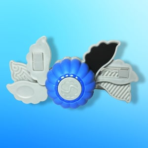 以下が含まれることがあります： 青とグレーの装飾品で、花のようなデザインです。中央は白いエンブレムが付いた青い円で、灰色の花びら型の要素に囲まれています。黒いアクセントが見えます。