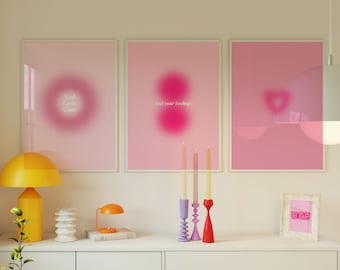 Peut inclure: Trois impressions d'art abstraites encadrées rose et blanc avec du texte qui dit "Sad Girls Club", "Feel your feelings" et une forme de cœur. Les impressions sont accrochées à un mur blanc au-dessus d'une commode blanche avec une lampe jaune, une petite lampe et des chandeliers.