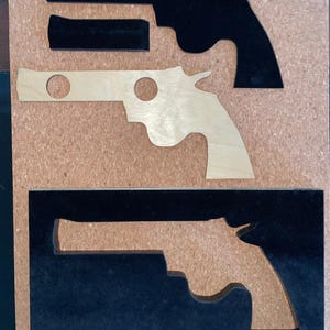 May include: A wooden puzzle featuring the outline of a revolver. The puzzle includes black and natural wood pieces, with the gun's silhouette cut out of the wood. The puzzle is set against a corkboard background.