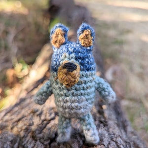 May include: A small crocheted stuffed animal in the shape of a bear. The bear is blue and grey with brown ears and a black nose.