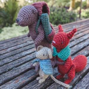 May include: Three crocheted stuffed animals: a brown bear with a green and brown backpack, a red squirrel with a teal scarf, and a small brown mouse with a blue scarf.