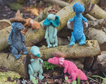 May include: A collection of six crocheted animal toys, including a blue dinosaur, a pink dinosaur, a green dinosaur, a brown and white dinosaur, a blue and white dinosaur, and a gray and white dinosaur. The toys are all sitting on a wooden surface.