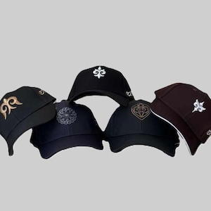 Puede incluir: Una colección de gorras de béisbol en varios colores, incluyendo negro y marrón. Cada gorra presenta un diseño bordado en oro o blanco. Las gorras están dispuestas en grupo, mostrando los diferentes estilos y colores disponibles.