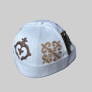 Puede incluir: Gorra blanca con patrones decorativos bordados en marrón y dorado. La gorra presenta una parte superior pequeña y redonda y un borde estructurado. El bordado incluye diseños florales y geométricos.