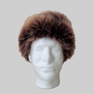 Può includere: Un cappello in finta pelliccia marrone è posto su una testa di manichino bianca. Il cappello ha una forma arrotondata e una consistenza soffice. La pelliccia è un mix di tonalità marroni chiare e scure. Accessorio invernale.