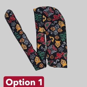 Puede incluir: Durag negro con un colorido estampado floral en rojo, verde y dorado. El durag está hecho de un material suave y tiene una cola larga. El texto "Opción 1" se muestra en un rectángulo rojo.