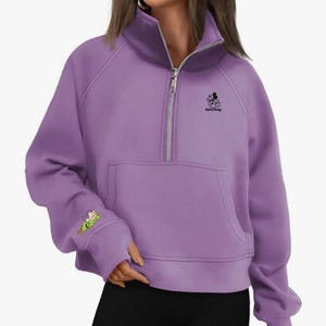 May include: A lavender-colored sweatshirt with a high collar and a silver zipper. The sweatshirt features a front pocket and a small graphic of Mickey Mouse with the text "runDisney". A cartoon character is on the left sleeve.