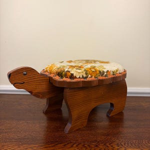 May include: A wooden footstool shaped like a turtle with a dark brown stained finish. The seat features a cushioned top upholstered in a cream fabric with a floral pattern in shades of orange, yellow, and brown, secured with decorative trim.