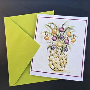 May include: Watercolor greeting card featuring a pineapple decorated with ornaments in shades of gold, purple, and silver. The card has a white background with a purple border and is paired with a bright green envelope.