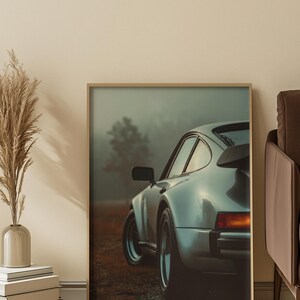 May include: Framed print of a silver sports car, partially visible, with a black spoiler. The car is parked on a gravel road, with a blurred, foggy background. The print is displayed in a room with a neutral color palette.