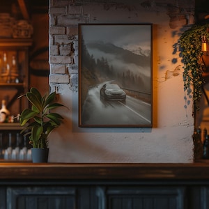 May include: A framed print depicting a car driving on a wet road through a mountain pass. The artwork is in a dark, moody style, with a focus on the car and the misty landscape. The frame is brown, and the print is hanging on a brick wall.