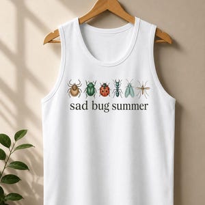 Peut inclure: Un débardeur blanc sans manches orné d'une rangée horizontale de six illustrations détaillées d'insectes, dont une tique, un scarabée, une coccinelle, une fourmi, une mouche et un moustique, avec le texte noir "sad bug summer" imprimé en dessous.