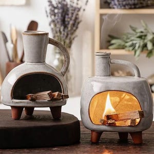 May include: Two light gray ceramic incense burners shaped like small fireplaces with handles and three terracotta-colored legs. One burner contains small wooden sticks, while the other features a lit flame and smoke rising from the top chimney.