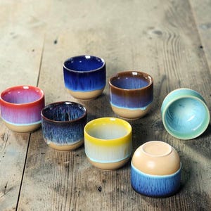 May include: A collection of seven ceramic cups featuring a two-tone reactive glaze. Each cup has a smooth, unglazed tan base and a vibrant, dripping colored upper section in shades of red, blue, brown, yellow, and light green, arranged together.