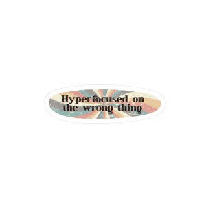 May include: An oval-shaped sticker featuring a distressed, retro-inspired sunburst pattern in muted shades of blue, red, yellow, and cream. The text "Hyperfocused on the wrong thing" is printed in a bold, dark, serif font across the center.