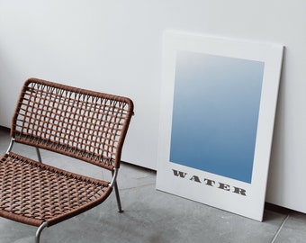 Op de afbeelding: Een bruine rieten stoel met een metalen frame staat voor een wit ingelijst poster met een blauwe gradiënt en het woord "WATER" in zwart.