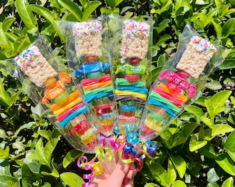May include: Four colorful candy skewers wrapped in clear cellophane with rainbow-colored gummy candies, white chocolate covered marshmallows, and colorful sprinkles. The skewers are tied with colorful ribbons.