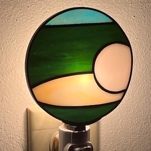 May include: A stained glass night light with a green, blue, and white design. The light is plugged into an outlet.