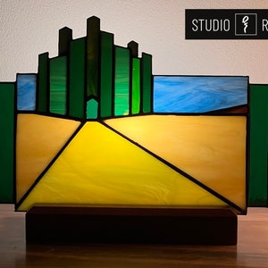 May include: A stained glass lamp depicting the yellow brick road leading to the Emerald City from the Wizard of Oz. The lamp is on a wooden base and features a blue sky above the road.