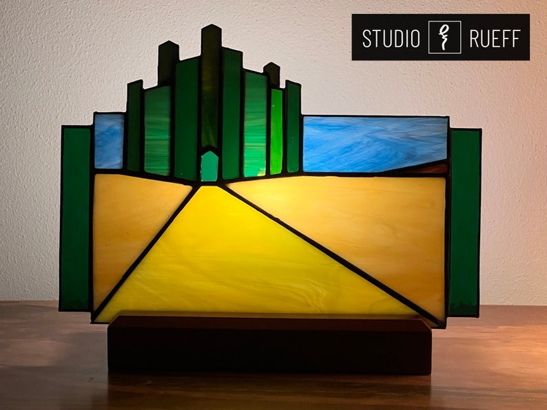 May include: A stained glass lamp depicting the yellow brick road leading to the Emerald City from the Wizard of Oz. The lamp is on a wooden base and features a blue sky above the road.