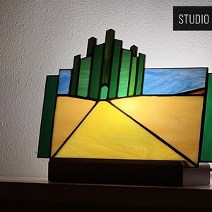 May include: A stained glass lamp featuring a yellow brick road leading to the Emerald City, a green cityscape with a door in the center. The lamp is on a wooden base and has a cord.