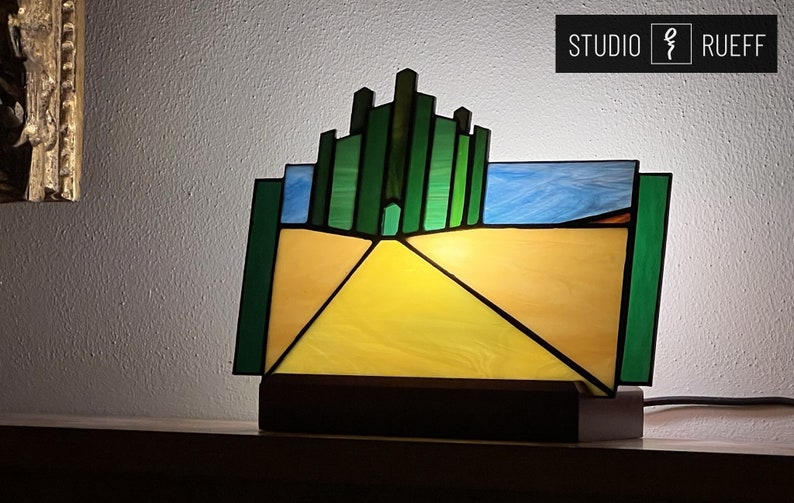 May include: A stained glass lamp featuring a yellow brick road leading to the Emerald City, a green cityscape with a door in the center. The lamp is on a wooden base and has a cord.