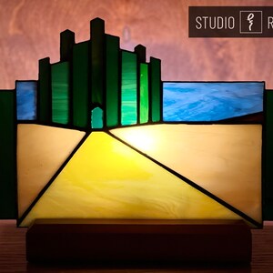 May include: A stained glass lamp depicting the Emerald City from the Wizard of Oz. The lamp features a yellow brick road leading to the city, which is made of green glass blocks. The lamp is lit from within, casting a warm glow.