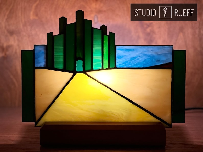 May include: A stained glass lamp depicting the Emerald City from the Wizard of Oz. The lamp features a yellow brick road leading to the city, which is made of green glass blocks. The lamp is lit from within, casting a warm glow.