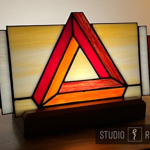 May include: A stained glass lamp with a geometric design featuring a red, orange, and yellow triangle. The lamp is on a wooden base and has a warm glow.