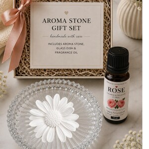 May include: An aroma stone gift set in a box with a pink ribbon, containing a white daisy-shaped aroma stone, a clear textured glass dish, and a 10 ml bottle of rose fragrance oil. The box label reads AROMA STONE GIFT SET, handmade with care.