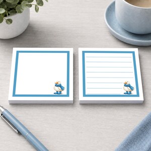 May include: Two square sticky note pads with a white centre and a blue border. Each pad features a small illustration of a white duck wearing a blue scarf and blue rain boots in the bottom right corner. One pad is blank, and one is lined.