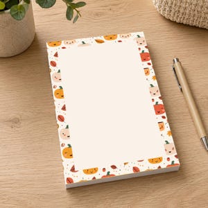May include: A rectangular notepad featuring a wide, cream-coloured central writing area bordered by a repeating pattern of small, smiling orange and light pink pumpkins, brown and orange autumn leaves, and tiny black dots on a white background.