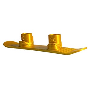 Puede incluir: Un snowboard decorativo en miniatura de color dorado con dos candelabros a juego en forma de bota. El snowboard tiene una superficie texturizada y una punta curvada. Las botas tienen un diseño de X en la parte delantera.