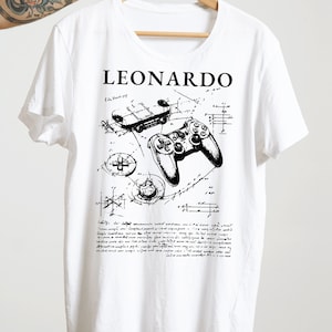 Puede incluir: Camiseta blanca con una ilustración en blanco y negro de un mando de videojuego y el texto "LEONARDO" en la parte delantera.